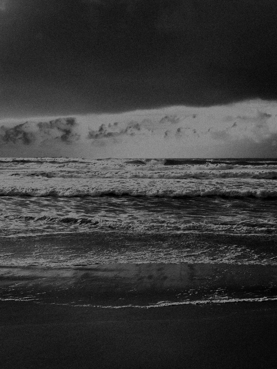 Photographie artistique en noir et blanc d’un paysage cotier désert, avec ciel nuageux et plage balayee par le vent. Style granuleux et minimaliste, evoquant la solitude, le silence et la poesie du littoral.