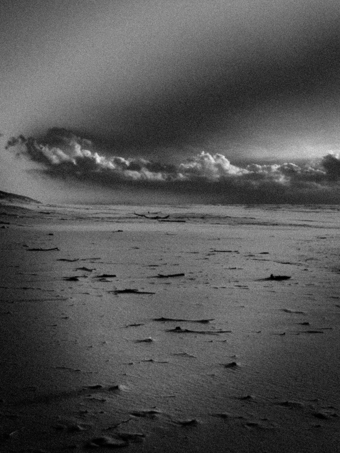 Photographie artistique en noir et blanc d’un paysage cotier désert, avec ciel nuageux et plage balayee par le vent. Style granuleux et minimaliste, evoquant la solitude, le silence et la poesie du littoral.